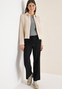 Beige textuur cardigan, grijze geribde top, zwarte wijde broek en licht olijfgroene sneakers. Sta tegen een witte muur.