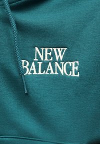 Tissu bleu sarcelle du sweat à capuche avec logo "NEW BALANCE" brodé en blanc et cordon bleu sarcelle assorti visible sur le côté gauche.