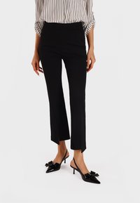 Pantaloni neri a vita alta e taglio corto con spacchi laterali all'orlo, abbinati a décolleté neri lucidi con punta a contrasto e dettagli a fiocco.