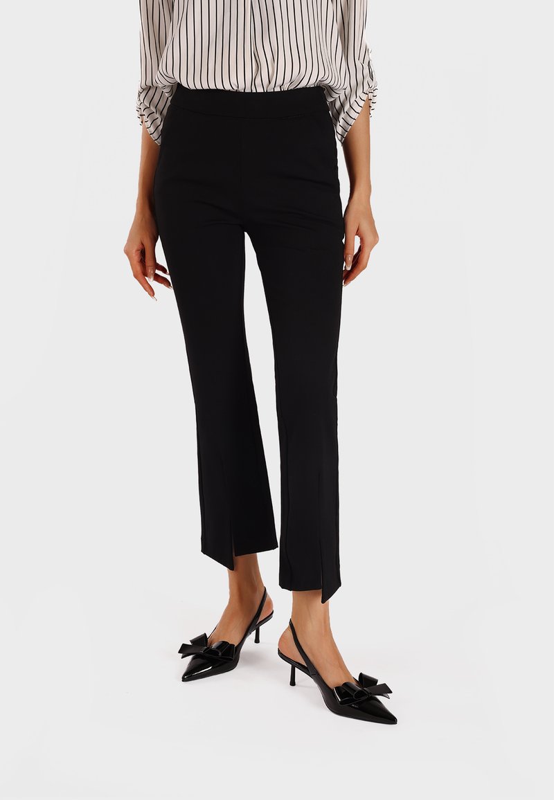 Pantaloni neri a vita alta e taglio corto con spacchi laterali all'orlo, abbinati a décolleté neri lucidi con punta a contrasto e dettagli a fiocco.
