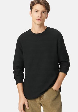 Joven con cabello castaño claro, vistiendo una camisa negra de manga larga texturizada y pantalones beige, de pie con una mano en el bolsillo contra un fondo blanco.