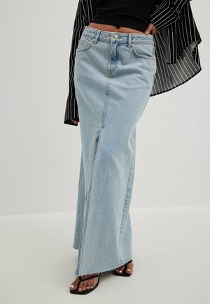 Vrouw draagt een lichtblauwe denim maxi-rok met ruwe zoom, zwarte sandalen met bandjes en een zwart gestreept cropped topje.