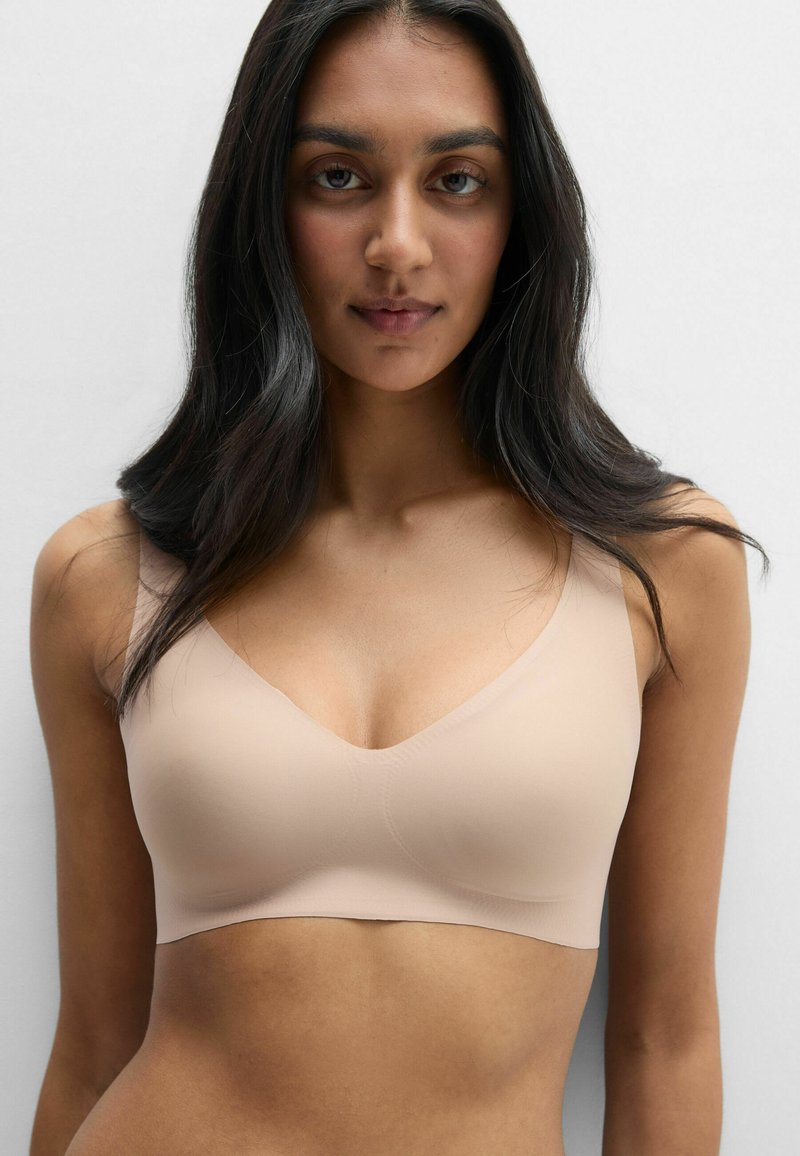 Kobieta z długimi ciemnymi włosami, ubrana w bezszwowy, cielisty biustonosz, zwrócona twarzą do przodu na tle jednolitego białego tła.