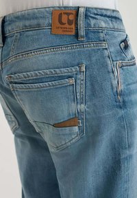 Jeans de mezclilla azul claro con una etiqueta de cuero que dice "LT" en la cinturilla. Presentan un bolsillo trasero con un acento marrón y una textura desgastada.