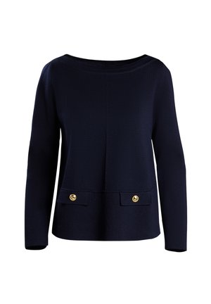 Frau trägt schwarzen Pullover über weißem Hemd mit Kragen, weiße weite Hosen, beige Ballerinas, hält weiße Handtasche mit schwarzem Schal.