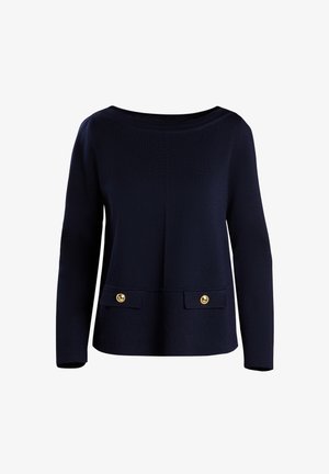 Femme portant un pull noir par-dessus une chemise blanche à col, pantalon blanc à larges jambes, chaussures plates beiges, tenant un sac à main blanc avec une écharpe noire.