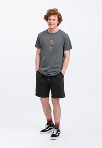 Grijze T-shirt met een kleurrijk grafisch ontwerp, gecombineerd met zwarte shorts. Model draagt zwarte sneakers met witte accenten en multicolore sokken.