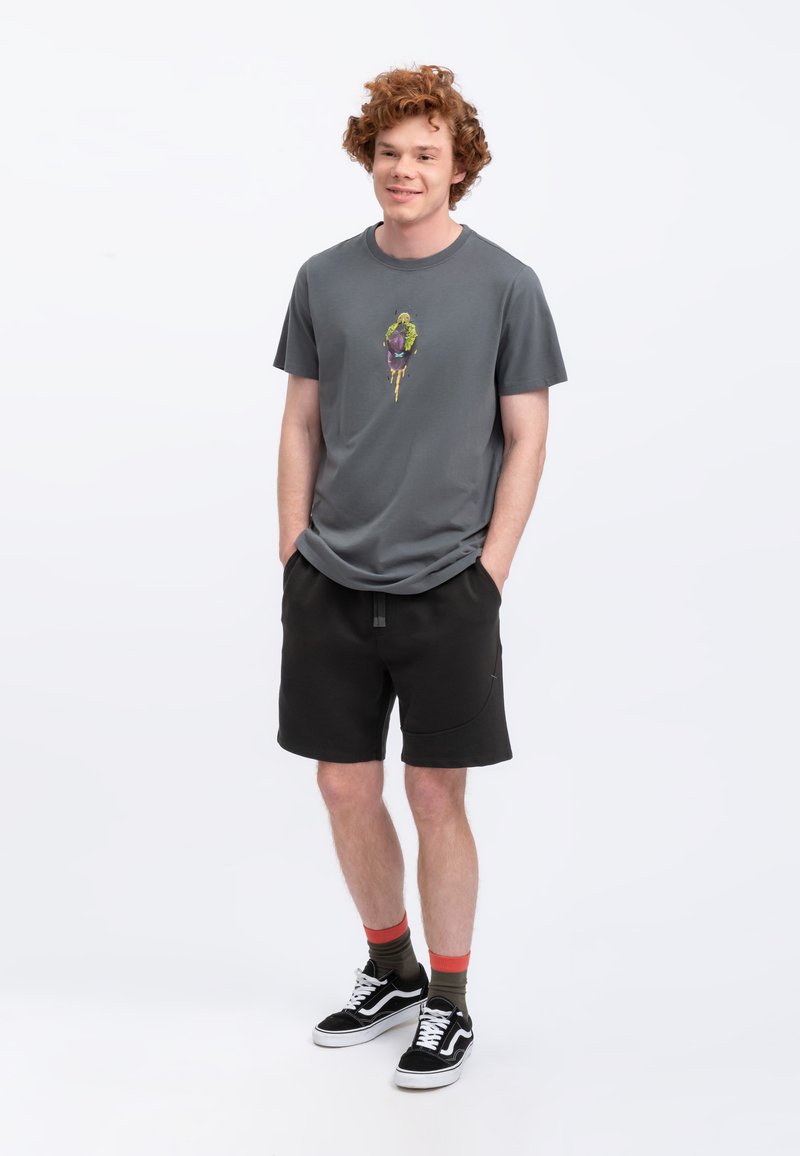 Grijze T-shirt met een kleurrijk grafisch ontwerp, gecombineerd met zwarte shorts. Model draagt zwarte sneakers met witte accenten en multicolore sokken.