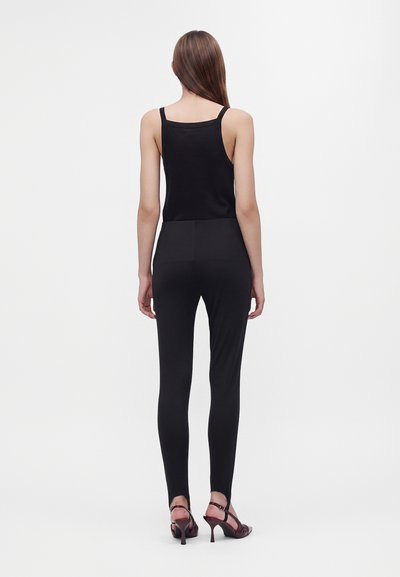 Femme de dos, portant un haut noir sans manches, un pantalon noir ajusté et des sandales à talons marron foncé sur un fond blanc uni.