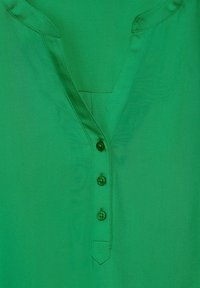 Blusa verde con scollatura a V, dotata di una patta con bottoni e cinque bottoni coordinati. Tessuto liscio e leggermente lucido, dettagli di cucitura minimi.