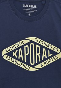 Námořnická modrá bavlněná trička s velkým krémovým diamantovým logem, na kterém je text "KAPORAL," "AUTHENTIC CLOTHING CO." a "ESTABLISHED."