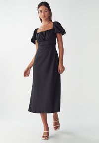 Musta, puff-hihainen midi-mekko, jossa on neliön muotoinen pääntie, rypistytty bodice ja sileä kangas. Yhdistetty valkoisiin remmisandaaleihin.
