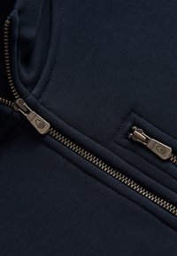 Sudadera navy con detalle de doble cremallera, que presenta una textura suave y un cuello acanalado. Cremalleras de metal expuestas con un brillo sutil.