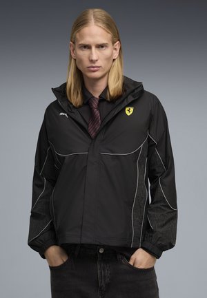 Person mit langen blonden Haaren trägt eine schwarze Puma- und Ferrari-Jacke über einem schwarzen Hemd und gestreifter Krawatte, die Hände in den Taschen schwarzer Jeans.