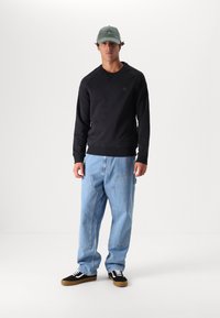 Felpa nera con scollo tondo, jeans di denim blu larghi e sneakers nere con lacci bianchi. Un cappellino grigio completa il look informale.
