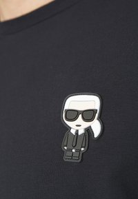T-shirt nera con una patch in gomma a forma di cartone animato che rappresenta un personaggio con capelli bianchi e occhiali da sole, che indossa un completo nero con cravatta.