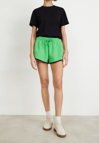 Gröna sportshorts med dragsko i midjan, svarta detaljer och en svart t-shirt. Matchas med vita sneakers och ljusa strumpor.