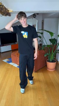 Fiatal férfi mosolyogva vakarja a fejét, fekete mintás pólót és cargo nadrágot visel, egy növényekkel és bútorokkal körülvett nappaliban áll.