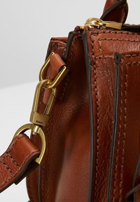 Coin d'un sac en cuir marron avec une couture détaillée et une boucle en métal doré reliant la sangle au sac.
