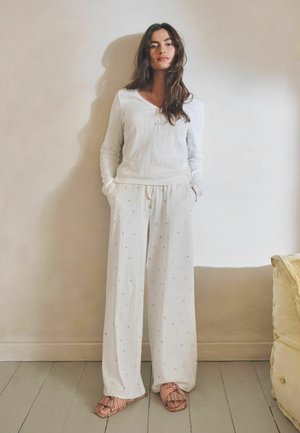 Femeie cu păr lung și închis la culoare, purtând un top alb cu mâneci lungi, pantaloni largi albi cu imprimeu floral mic și papuci deschiși cu dungi, stând în interior.