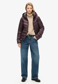 Veste rembourrée en violet foncé, dotée d'une capuche et d'un design matelassé. Portée sur un col roulé beige et associée à un jean bleu à jambes larges.