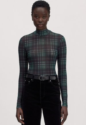 Vrouw draagt een aansluitende groen-zwarte ruit coltrui met lange mouwen, gestopt in een zwarte broek met hoge taille en een zwarte riem.