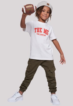 Ein Kind trägt ein weißes T-Shirt mit rotem Text, olivgrüne Cargo-Hosen mit elastischen Bündchen und weiße Turnschuhe, während es einen braunen Fußball hält.