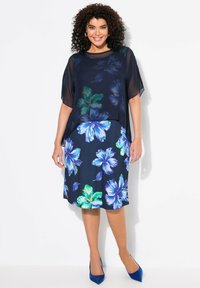 Námořnické květinové šaty s krátkými průhlednými rukávy, zdobené velkými modrými a zelenými květy. Doplňují je modré lodičky na vysokém podpatku.