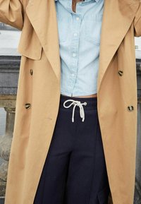 Trench-coat kaki à double boutonnage, larges revers et boutons foncés, porté sur une chemise bleue claire à boutons et un pantalon de survêtement bleu marine.