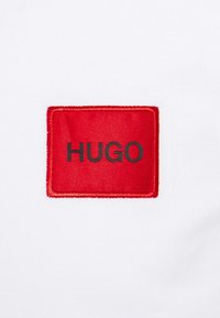 Un tissu blanc présente un patch rectangulaire rouge brodé avec le mot « HUGO » en noir. Le patch a une bordure texturée.