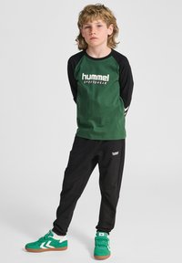 Maglietta verde a maniche lunghe con maniche nere e logo "hummel SPORTSWEAR", abbinata a pantaloni neri e sneakers verdi/bianche.