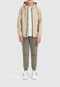 Licht beige lichtgewicht jack met capuchon, zwarte rits, gecombineerd met olijfgroene cargobroeken en witte sneakers met groene accenten, rechtopstaand.
