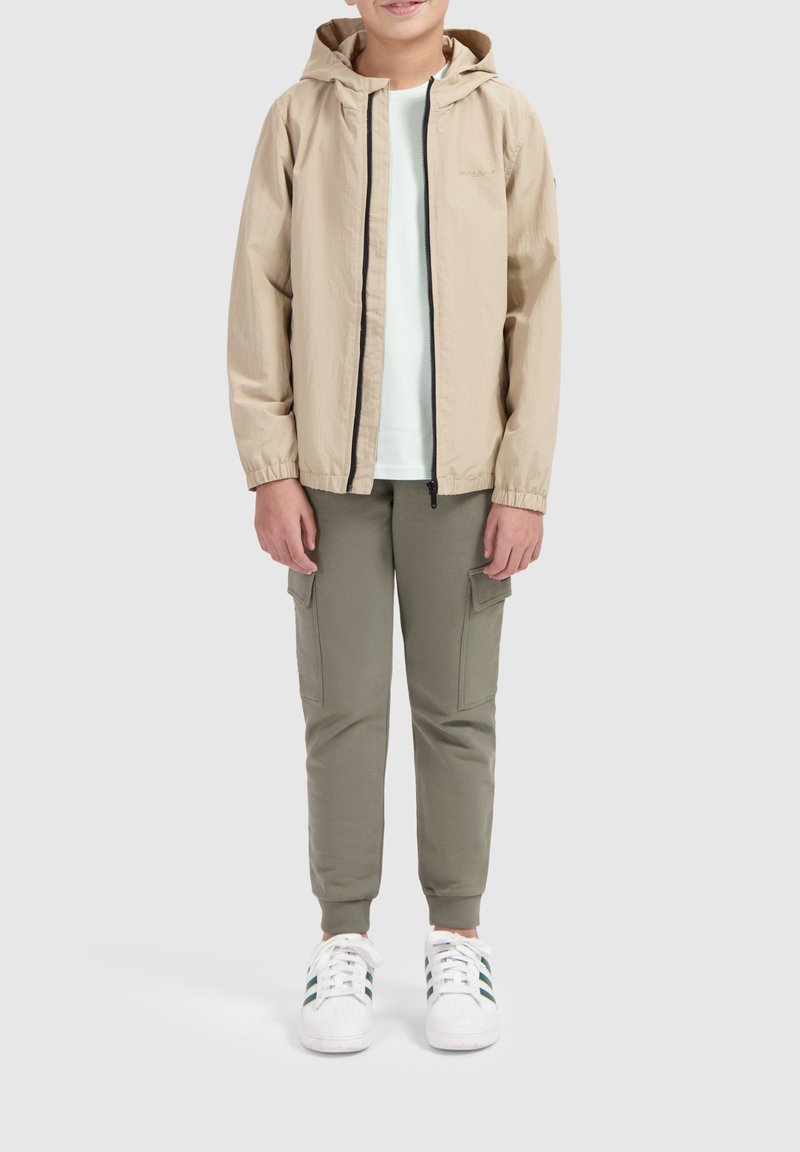 Licht beige lichtgewicht jack met capuchon, zwarte rits, gecombineerd met olijfgroene cargobroeken en witte sneakers met groene accenten, rechtopstaand.