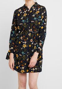 Kvinna klädd i en marinblå blommig långärmad skjortklänning med elastisk midja och krage, som står mot en enfärgad ljus bakgrund.