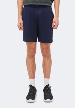 Homme portant un short de sport bleu marine, des chaussettes blanches et des chaussures de course noires, debout devant un fond clair uni.