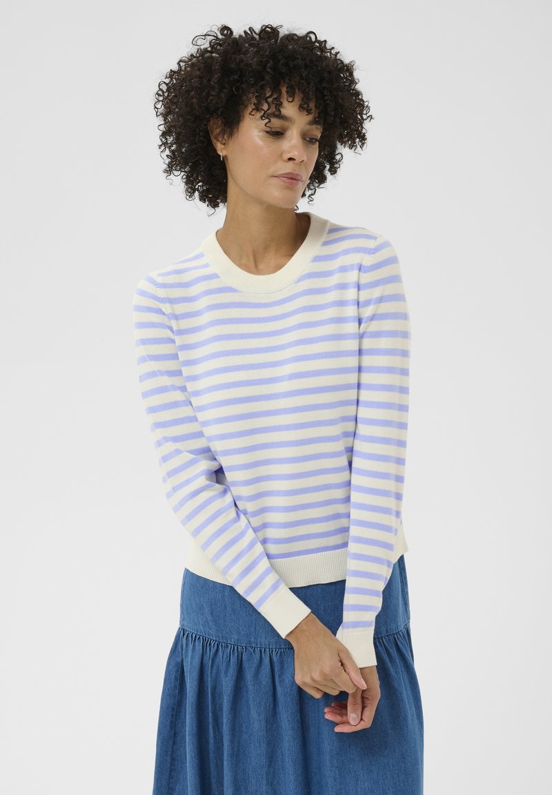 Femme aux cheveux bouclés portant un pull à manches longues blanc et bleu clair à rayures et une jupe bleue, debout devant un fond uni.