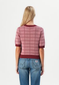 Femme blonde portant un pull à manches courtes à carreaux rose et bordeaux et un jean bleu avec des poches arrière cousues, se tenant de dos.