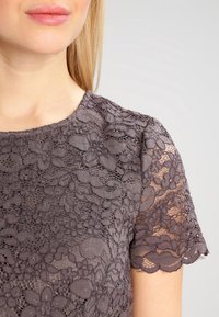 Szara koronkowa bluzka z floralnym wzorem, z krótkimi, falbaniastymi rękawami oraz okrągłym dekoltem. Widoczne są detale teksturowanej tkaniny.