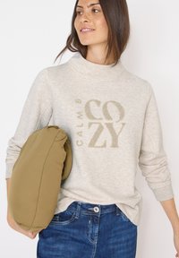 Bežová mikina s kovovým nápisem "CALM & COZY"; měkká tkanina, vysoký límec a dlouhé rukávy, v kombinaci se složenou olivově zelenou taškou.