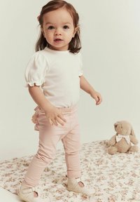 Haut blanc à manches courtes et côtes, leggings roses avec des détails en volants, et chaussures de couleur claire. Décor : couverture à motif floral avec un lapin en peluche.