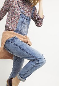 Salopette en denim bleu clair avec une texture délavée et des genoux abîmés, associée à un haut à motifs floraux et à un cardigan léger en tricot.