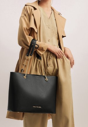 Femme portant un trench-coat beige et une tenue assortie, tenant un grand sac fourre-tout en cuir noir avec des anses en chaîne dorée.