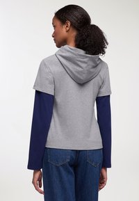 Felpa grigia a maniche corte con cappuccio, sovrapposta a una maglietta a maniche lunghe blu. Vestibilità casual, tessuto textured, indossata con jeans in denim blu.