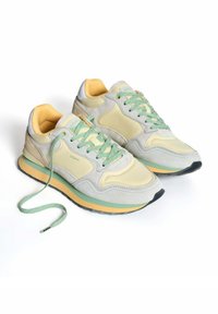 Zapatillas gris claro con acentos amarillos y verdes, materiales de ante y malla, y una suela de goma con patrón. Cordones en un verde a juego.