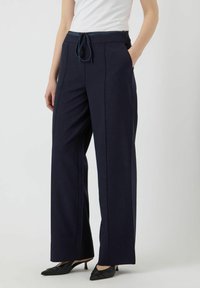 Pantalons larges bleu marine avec taille à cordon, poches latérales et texture lisse. Associés à des chaussures à talons noirs pour un look épuré.