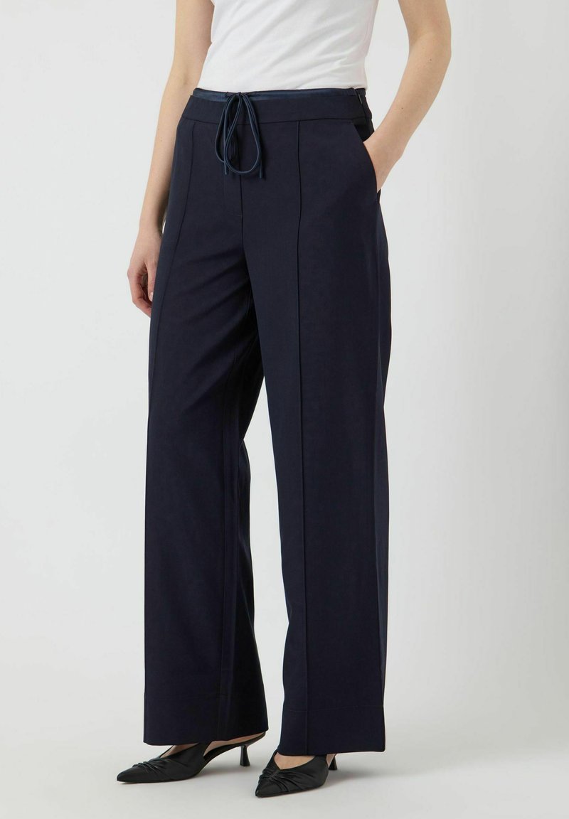 Pantalons larges bleu marine avec taille à cordon, poches latérales et texture lisse. Associés à des chaussures à talons noirs pour un look épuré.