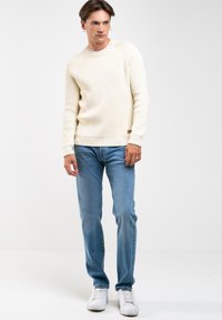 Suéter de punto acanalado en crema con cuello redondo, combinado con jeans rectos de lavado claro y zapatillas blancas. Etiqueta de marca sutil en el dobladillo del suéter.