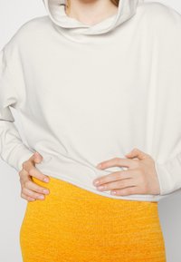 Felpa bianca oversize con cappuccio, abbinata a una gonna di maglia gialla texturizzata. Focus sui dettagli di design delle maniche e della vita.