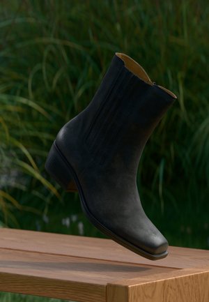 Bottine noire en cuir à petit talon suspendue au-dessus d'un banc en bois, avec un feuillage vert flou en arrière-plan.