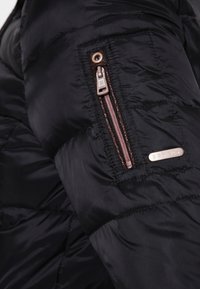 Svart puffjacka i närbild, med en dragkedjeficka på ärmen, glänsande nylonmaterial och kviltat mönster. Detaljer i roséguld.